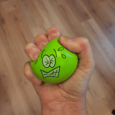 Een hand knijpt in een groene stressbal met een gespannen gezichtje, wat de spanning en druk symboliseert die mensen ervaren bij stressklachten en angsten.